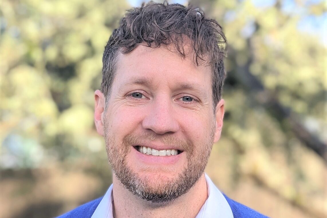 A man in blue shirt and white collared shirt smiling.